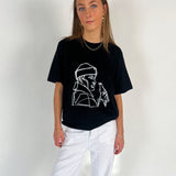En person iført en sort t-shirt med hvidt lineart-motiv og hvide bukser står i et lyst rum. Vedkommende har langt hår og bærer smykker.