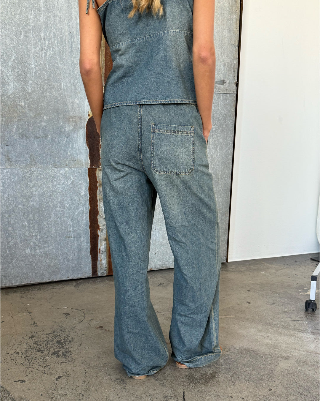 En person iført denim jumpsuit står med ryggen til, hænderne i lommerne. Baggrunden er en kombination af gråmetal og hvid væg på et betongulv.