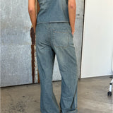 En person iført denim jumpsuit står med ryggen til, hænderne i lommerne. Baggrunden er en kombination af gråmetal og hvid væg på et betongulv.