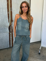 Kvinde i denim jumpsuit står afslappet med hænderne i lommerne, foran en rusten metalbaggrund og en hvid væg, med hård overgang i en fotostudiosetting.