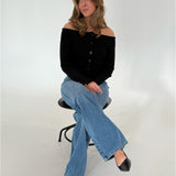 En kvinde sidder på en høj sort skammel. Hun bærer en sort off-shoulder bluse og blå jeans. Rummet omkring hende er hvidt og neutralt.