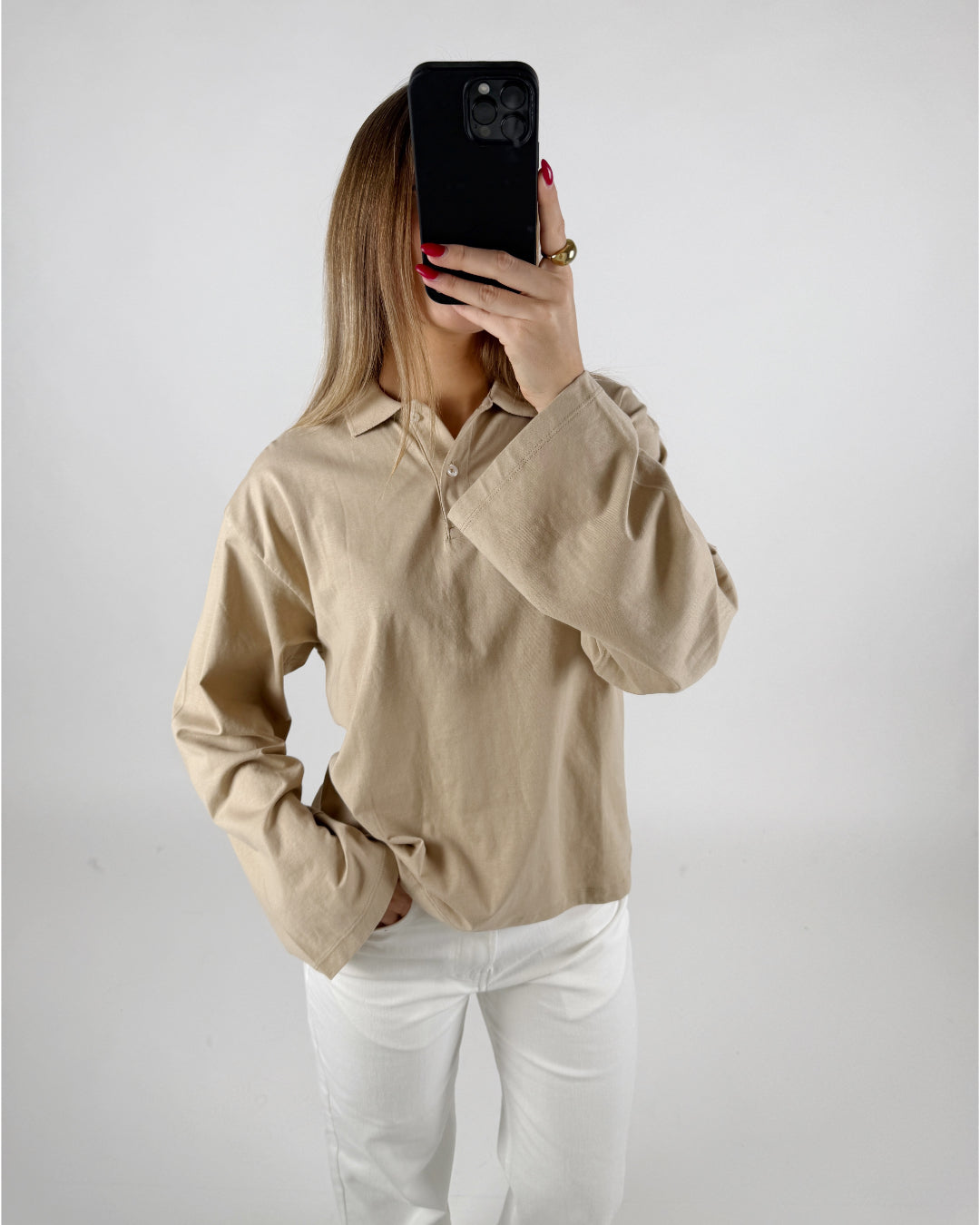 En person iført en beige skjorte og hvide bukser holder en sort smartphone foran ansigtet i et neutralt, lys baggrundsmiljø.