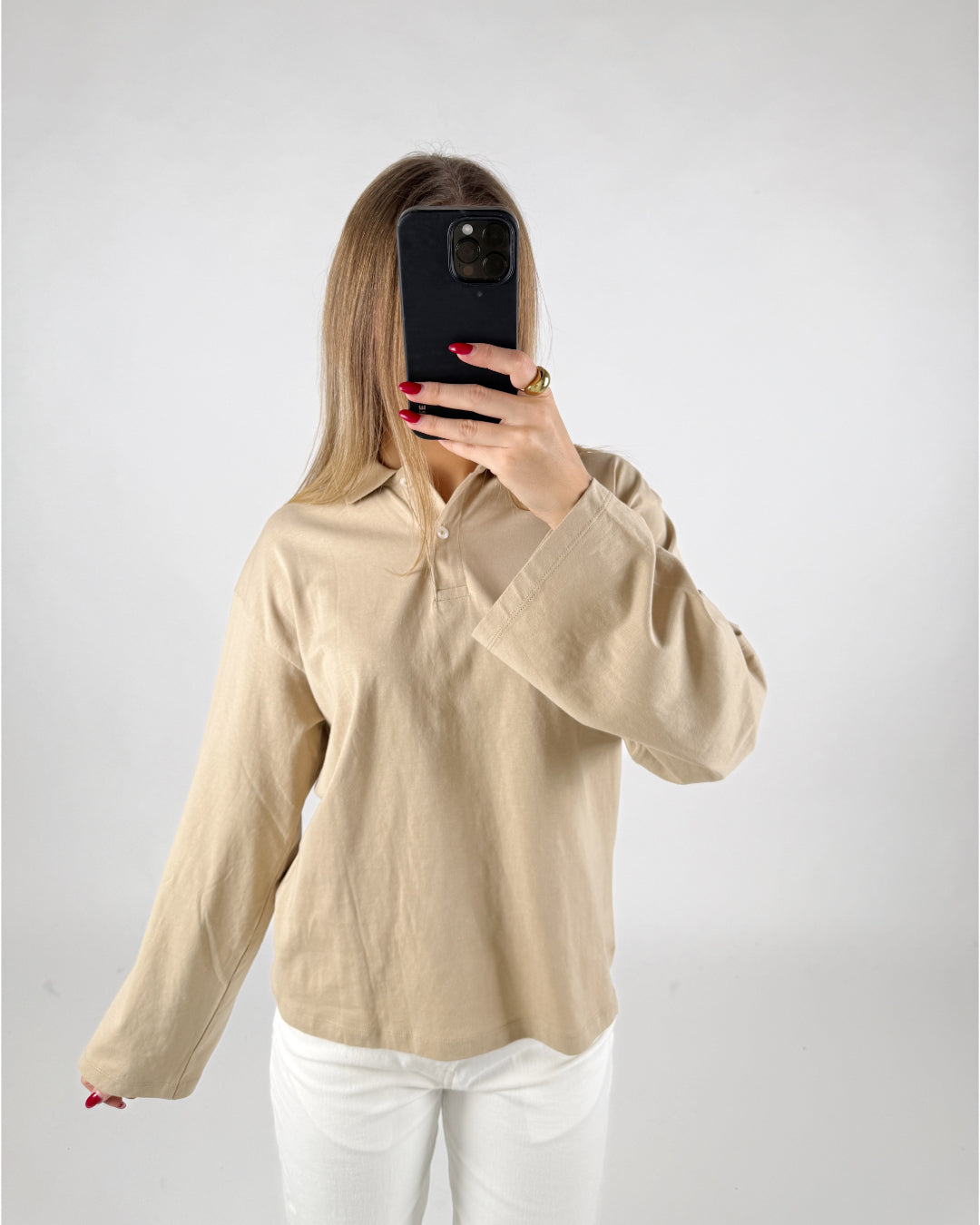 Person tager et billede med en smartphone; iført beige skjorte og hvide bukser. Personen er i et rum med en ensfarvet hvid baggrund.