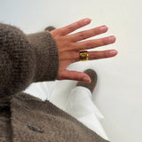 Hånd bærer en gylden ring og peger nedad. Personen har en brunt sweater og hvide bukser på. Støvler er synlige i baggrunden på en hvid baggrund.