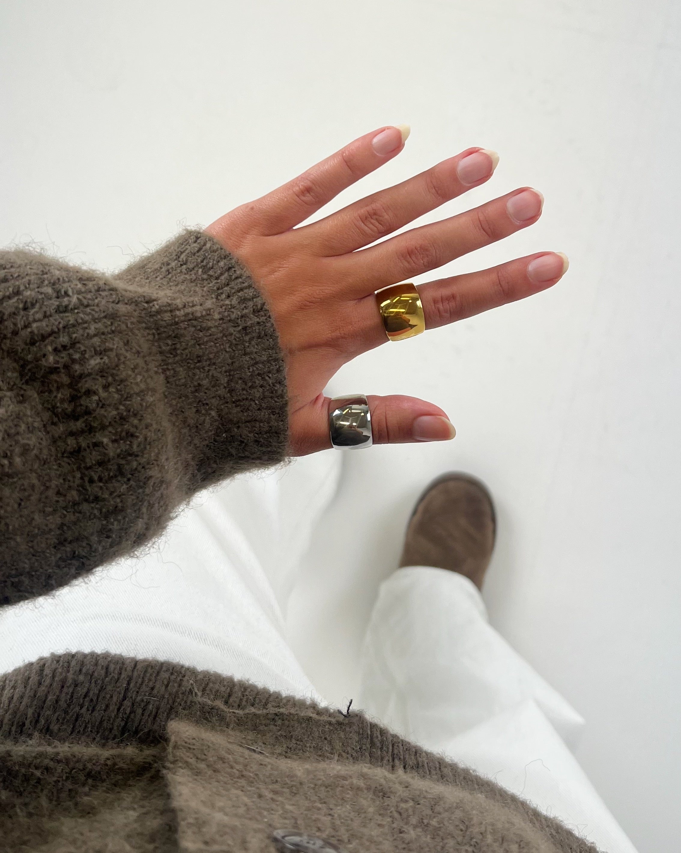 En hånd med en brun sweater bærer to store ringe, én guld og én sølv. Baggrunden viser hvide bukser og en brun sko på et lyst gulv.