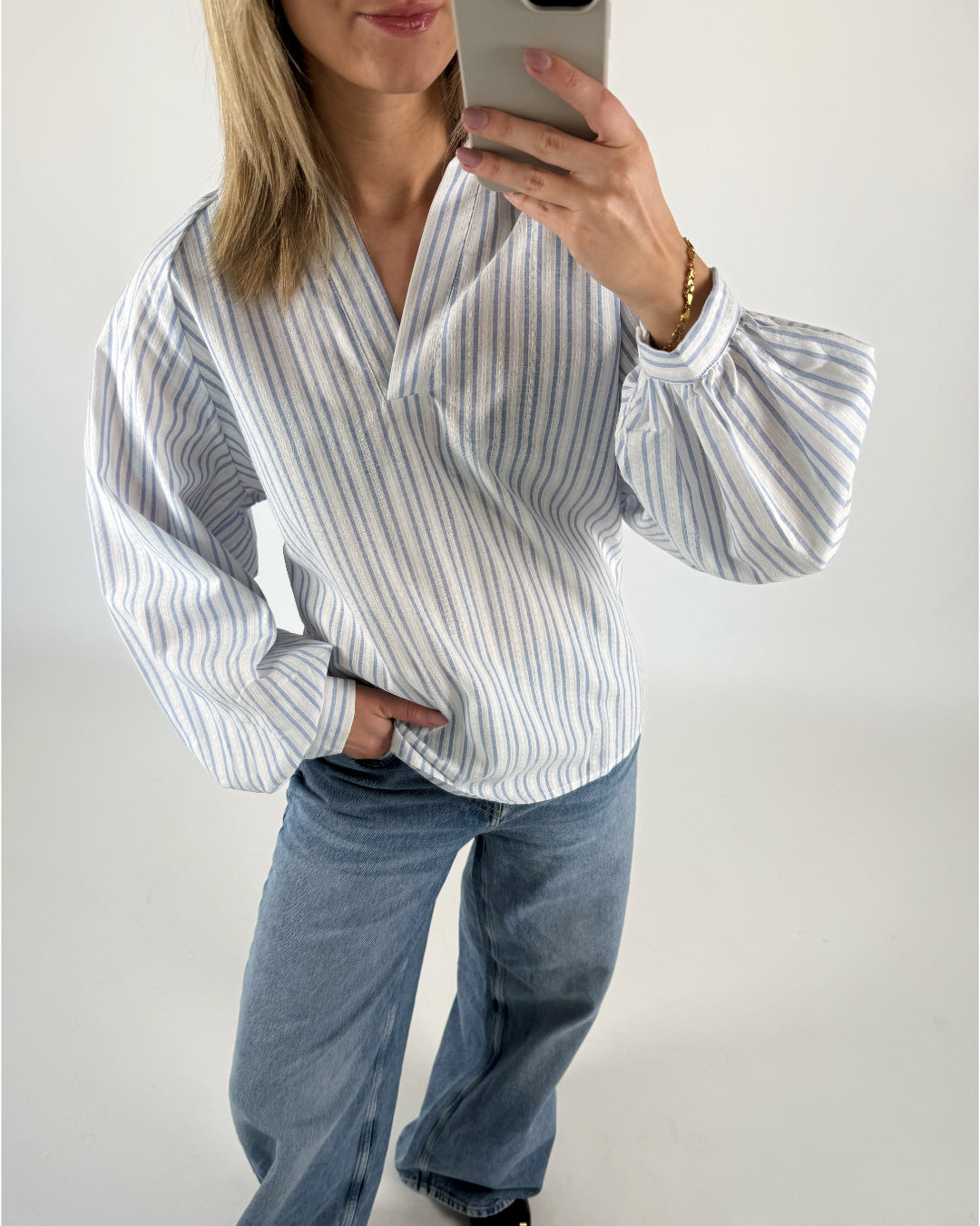 Kvinde iført stribet bluse tager selfie med telefon, står afslappet med en hånd i lommen i lyseblå jeans mod en hvid baggrund.