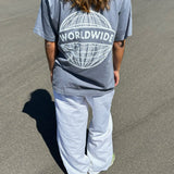 Person iført grå t-shirt med teksten “WORLDWIDE” over en globusgrafik, står på en asfaltvej under solen, iført hvide bukser.