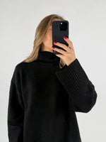 En kvinde i en sort rullekrave trøje holder en sort telefon op foran sit ansigt i en hvid baggrund.