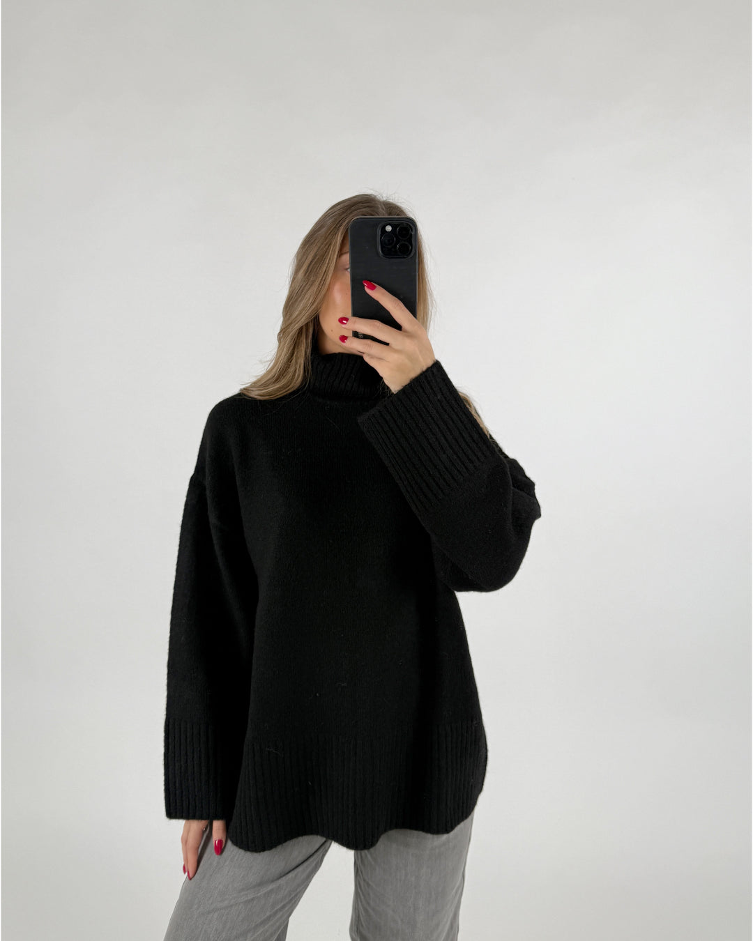 En kvinde holder en sort smartphone foran sit ansigt. Hun er iført en sort rullekrave sweater og grå bukser, i et hvidt studie.