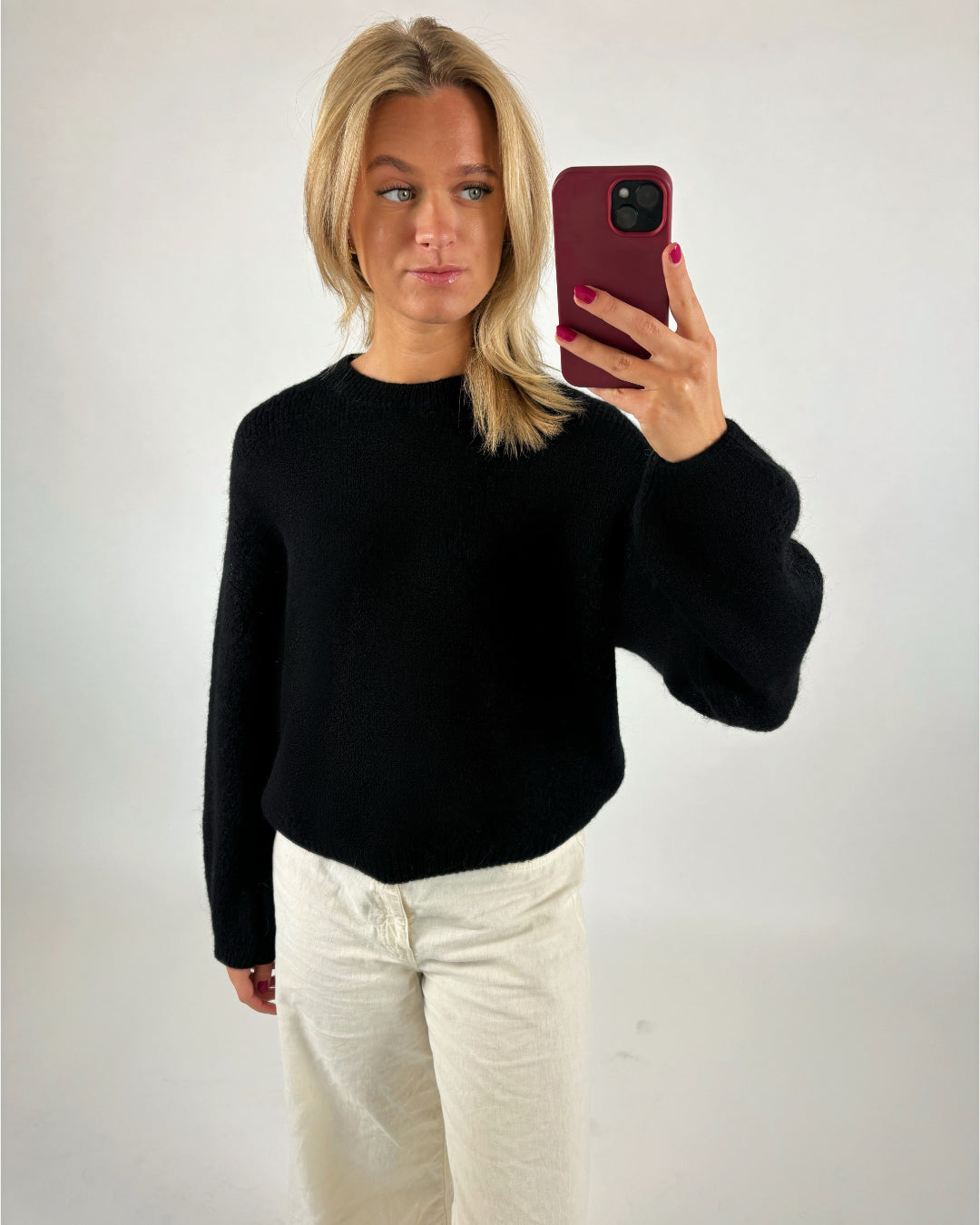 En person iført en sort trøje og hvide bukser tager en selfie med en rød telefon i et neutralt oplyst rum.