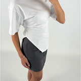 En person iført en hvid t-shirt og mørk nederdel holder en mobiltelefon, fotograferet fra siden i et hvidt studie. Hendes arm bærer to armbånd.