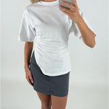 Kvinde iført hvid T-shirt og grå nederdel tager et spejl-selfie. Hun står i et hvidt rum og holder en smartphone med sølvfarvet etui.