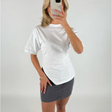 En person iført en hvid t-shirt og en mørkegrå nederdel holder en mobiltelefon foran ansigtet i et minimalistisk, lyst rum.