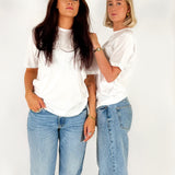 To kvinder står stille iført hvide T-shirts og blå jeans mod en enkel, hvid baggrund. Den ene har langt mørkt hår, den anden kort lyst hår.