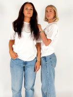 To kvinder står stille iført hvide T-shirts og blå jeans mod en enkel, hvid baggrund. Den ene har langt mørkt hår, den anden kort lyst hår.