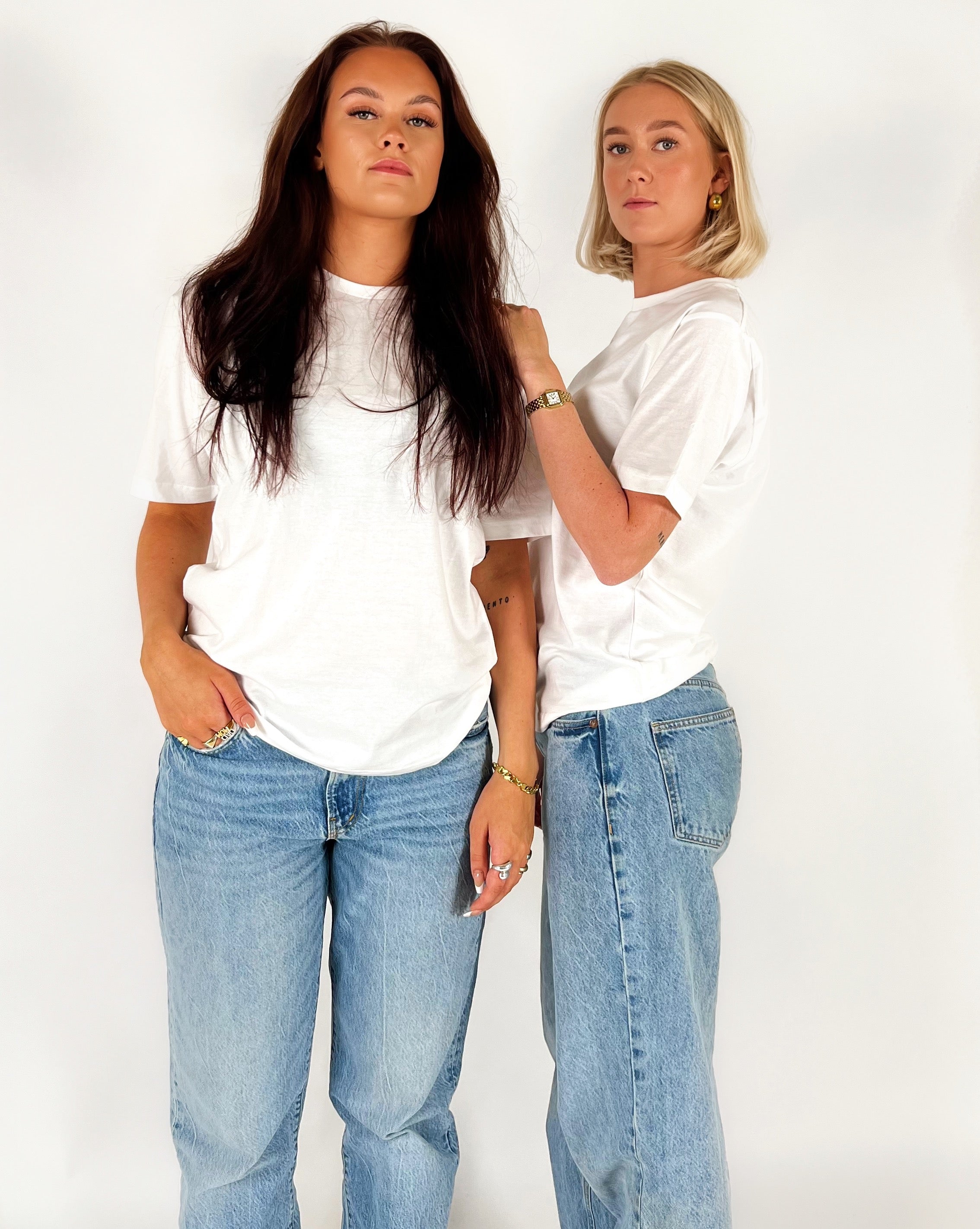 To kvinder står stille iført hvide T-shirts og blå jeans mod en enkel, hvid baggrund. Den ene har langt mørkt hår, den anden kort lyst hår.