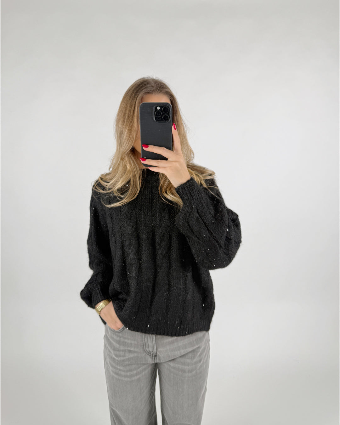 En person tager et billede med en smartphone i et spejlløst rum. De bærer en sort sweater og grå bukser, med hånden i lommen. Baggrunden er enkel og lys.