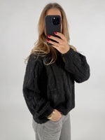 En person iført en sort striktrøje tager et billede med en sort smartphone. De har løst hår og røde negle. Baggrunden er enkel og lys. Ingen tekst til stede.