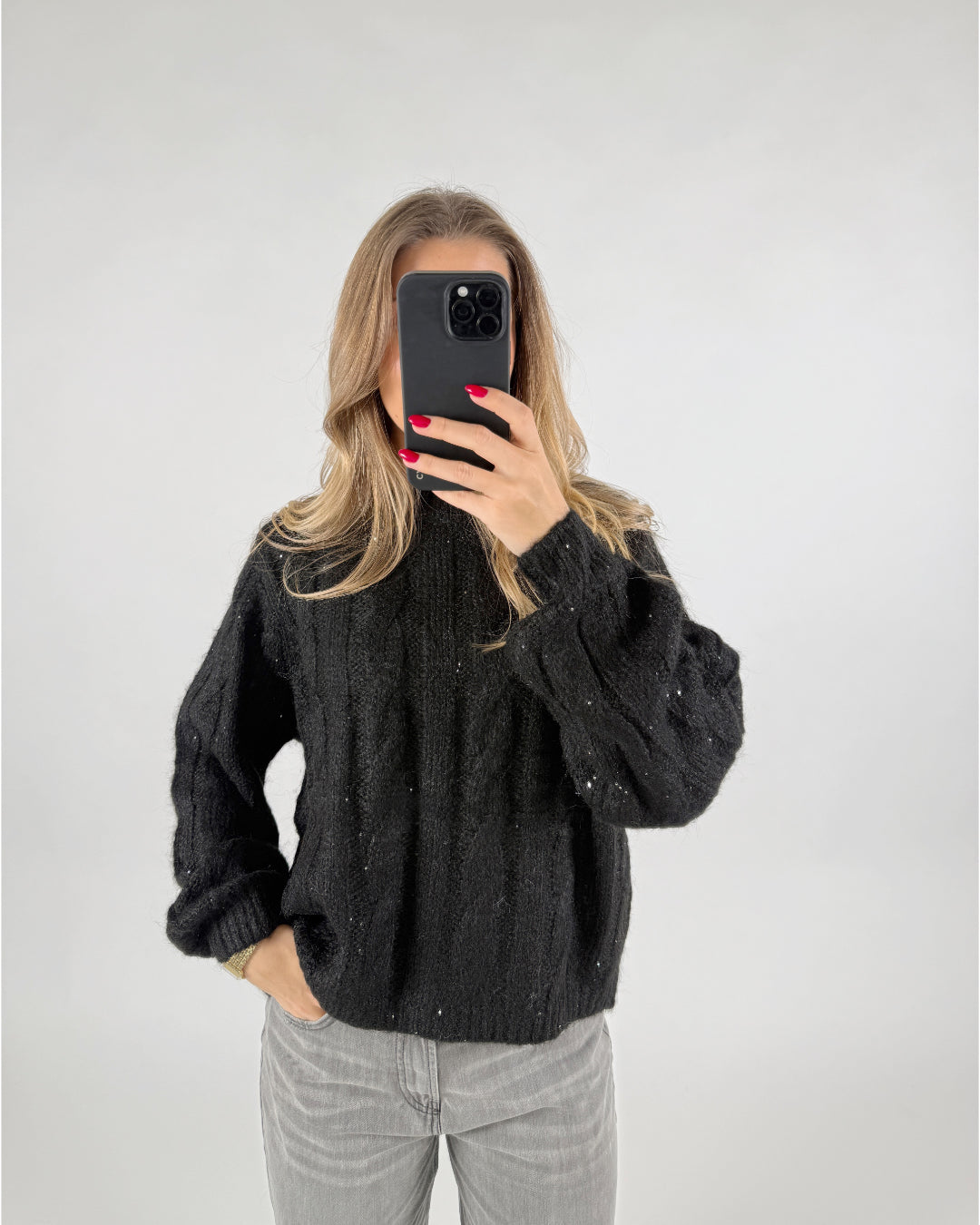 En person i en sort sweater holder en mobiltelefon foran ansigtet i et neutralt oplyst rum. De har én hånd i lommen på deres grå bukser.