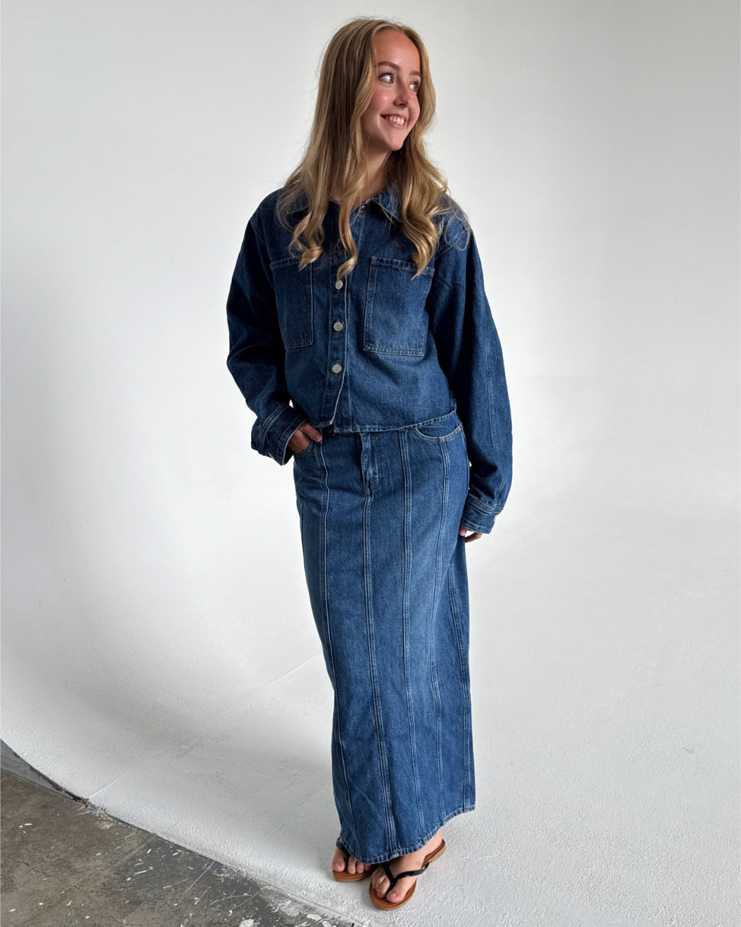 Modelfoto af pige som bærer en blå Cana denim nederdel, samt Cana denim jakke og sorte Carmen sandaler. Alt fra By Sofie Sønderby