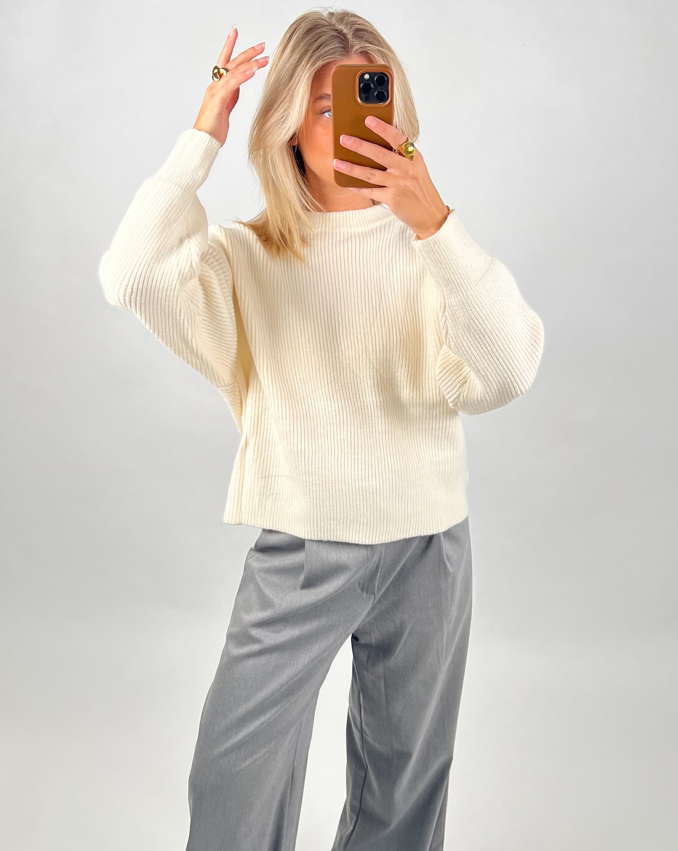 Person tager et billede. Iført en hvid sweater og grå bukser. De står foran en lys baggrund, og bruger en mobiltelefon med brunt etui til at tage billedet.