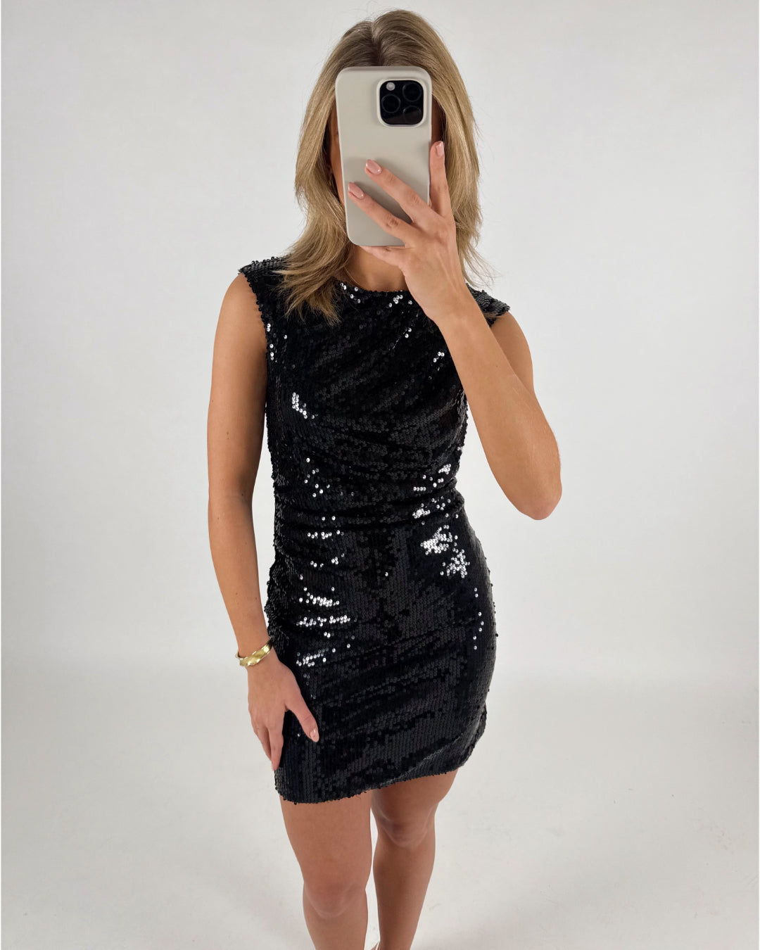 En person iført en sort, glitrende kjole tager en spejlselfie med en mobiltelefon. Rummet er enkelt med en lys baggrund.