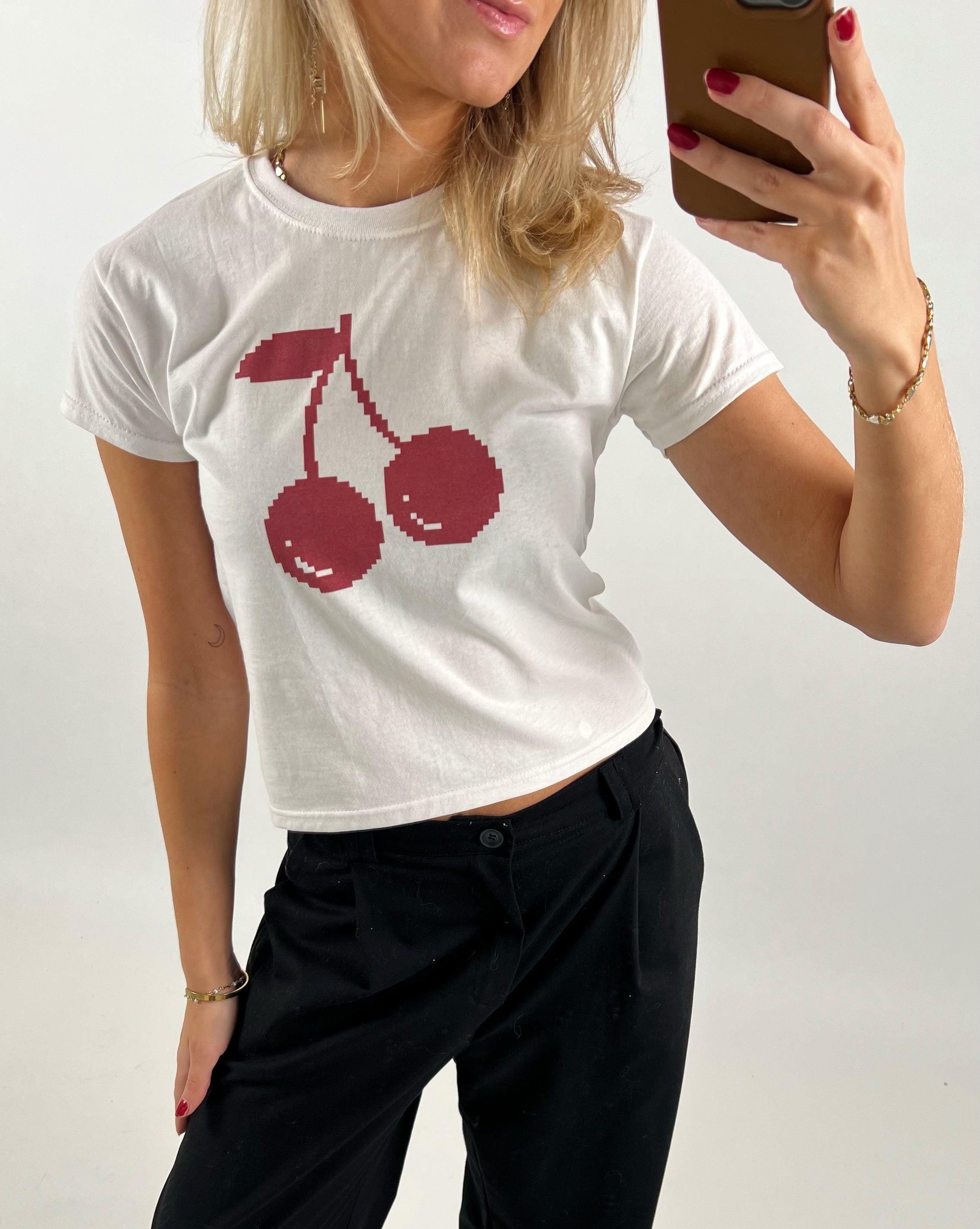 En person iført hvid t-shirt med rødt kirsebærmotiv tager en selfie i et spejl. Personen har sort bukser, rød neglelak, gyldent armbånd og holder en brun telefon.