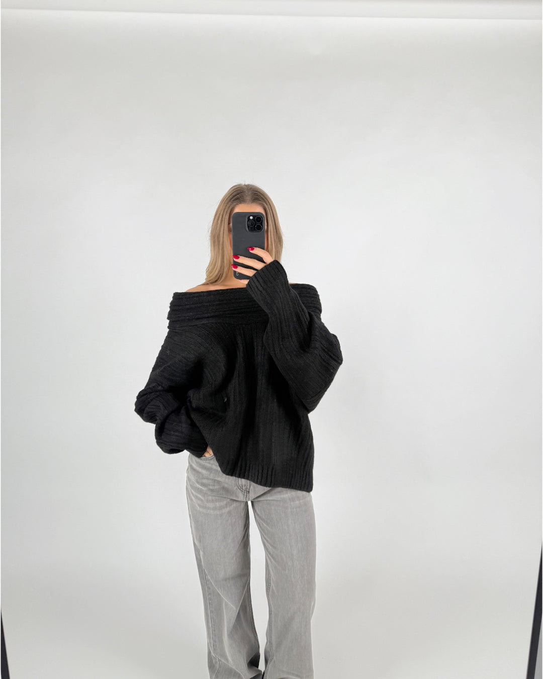 En person iført en sort trøje poserer med en hånd i lommen og en mobiltelefon i den anden, i et minimalistisk, lyst rum.