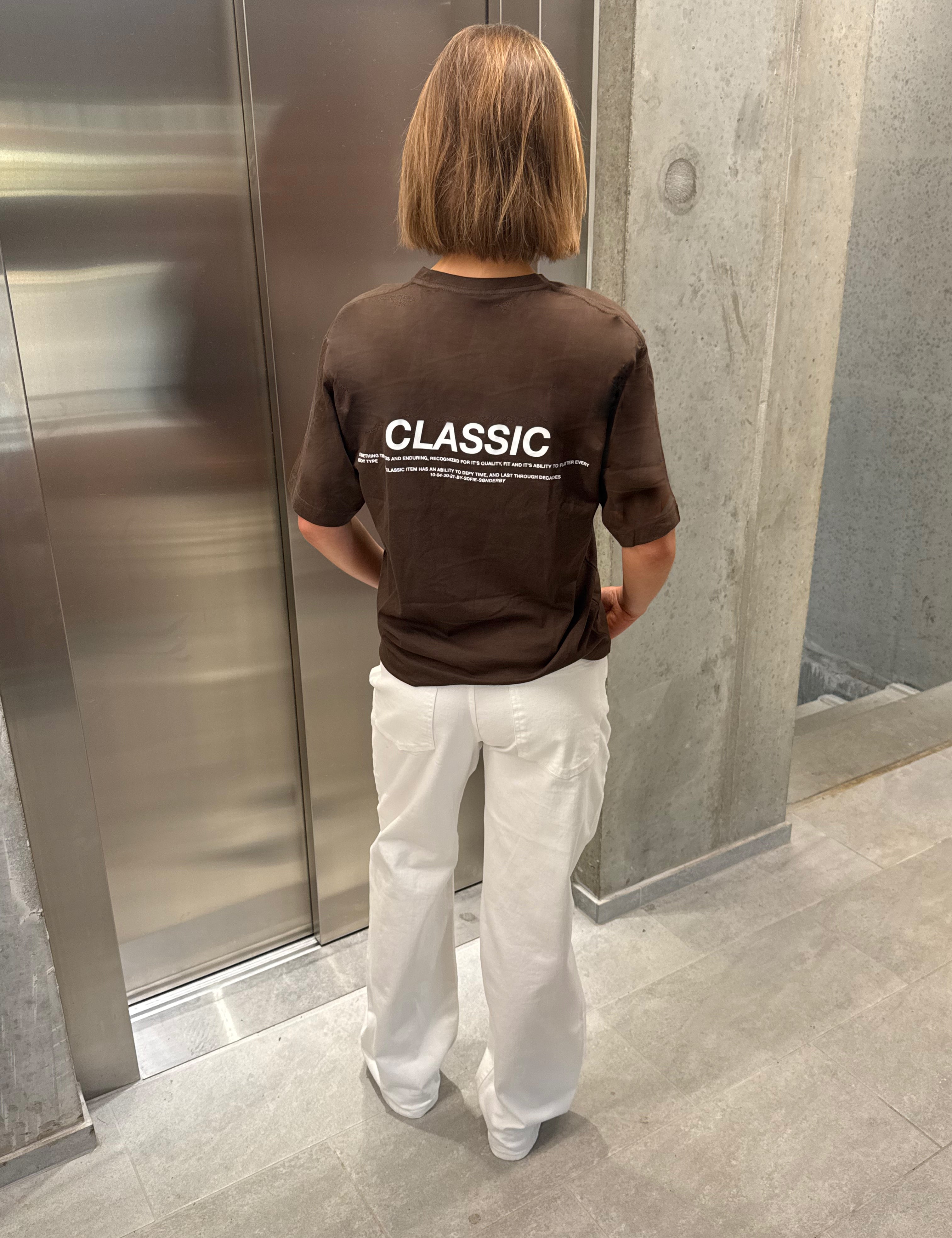 Stemningsbillede af pige der bærer en brun t-shirt med hvidt classic tryk og hvide NANNA jeans.  Fra BySofieSønderby