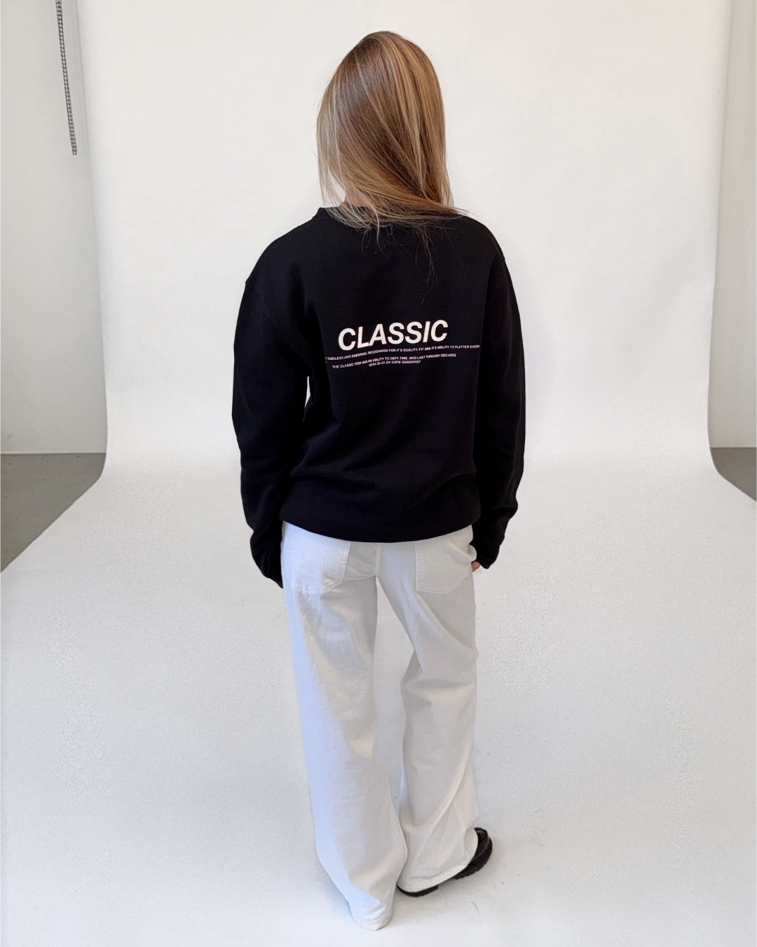 Modelfoto af pige i sort CLASSIC sweatshirt med reflekstryk, samt hvide NANNA jeans. Alt er fra BySofieSønderby.