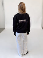 Modelfoto af pige i sort CLASSIC sweatshirt med reflekstryk, samt hvide NANNA jeans. Alt er fra BySofieSønderby.