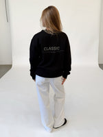 Modelfoto af pige i sort CLASSIC sweatshirt med reflekstryk, samt hvide NANNA jeans. Alt er fra BySofieSønderby.