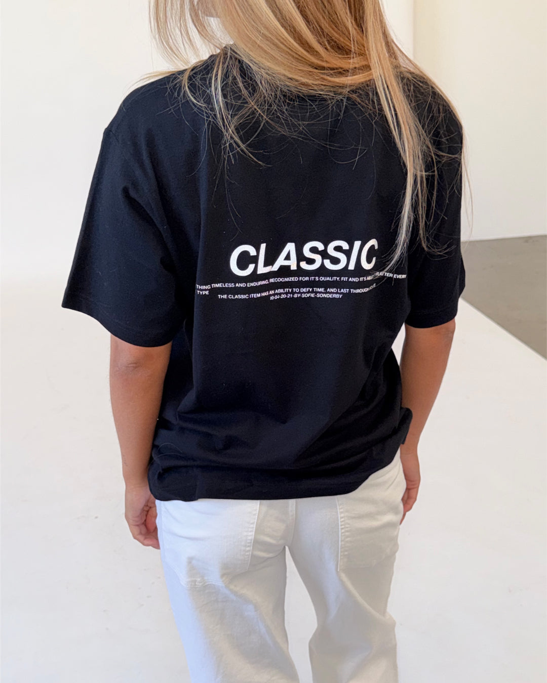 Modelfoto af pige i sort CLASSIC t-shirt med reflekstryk, samt hvide NANNA jeans. Alt er fra BySofieSønderby.