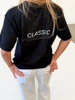 Modelfoto af pige i sort CLASSIC t-shirt med reflekstryk, samt hvide NANNA jeans. Alt er fra BySofieSønderby.