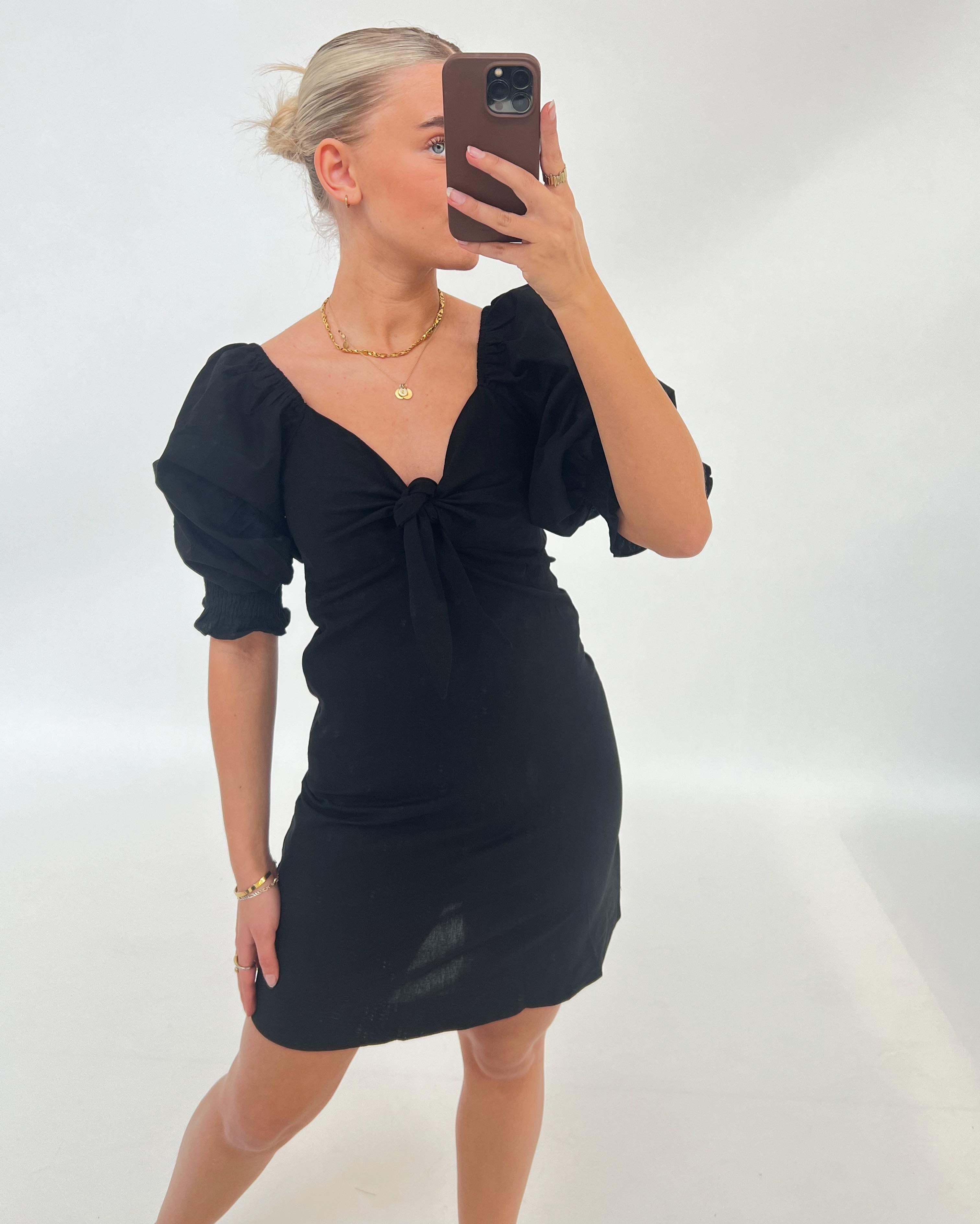 Kvinne i sort kjole tager selfie med sin mobiltelefon i et lyst rum. Hun har håret opsat og bærer guldsmykker.