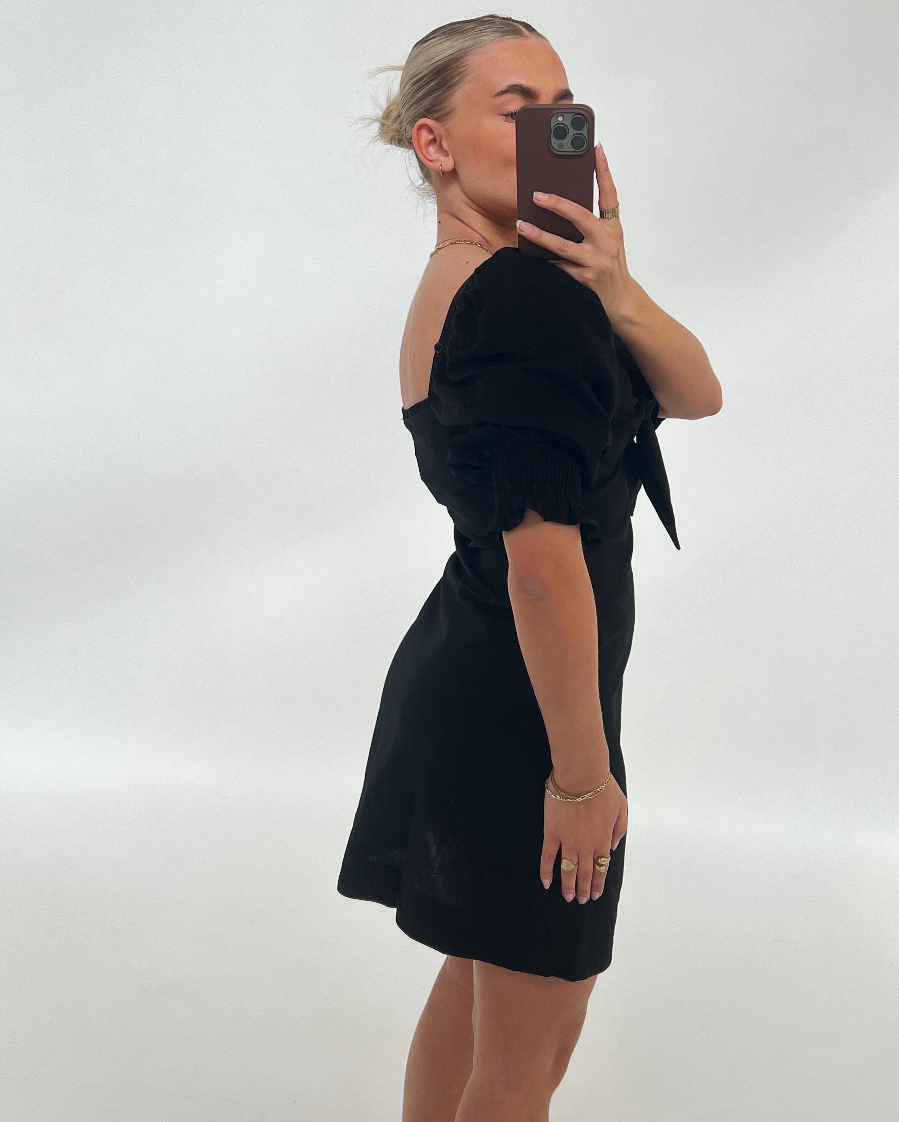 En person i sort kjole tager et spejlselfie med mobilen i en lys baggrund. Personen har lyst hår opsat i en knold og bærer smykker.