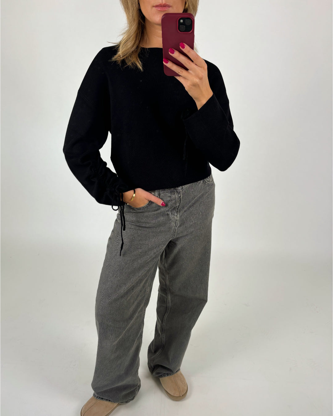 En person holder en telefon i et selfie, iført en sort trøje og grå bukser. Personen står foran en ensfarvet, lys baggrund.