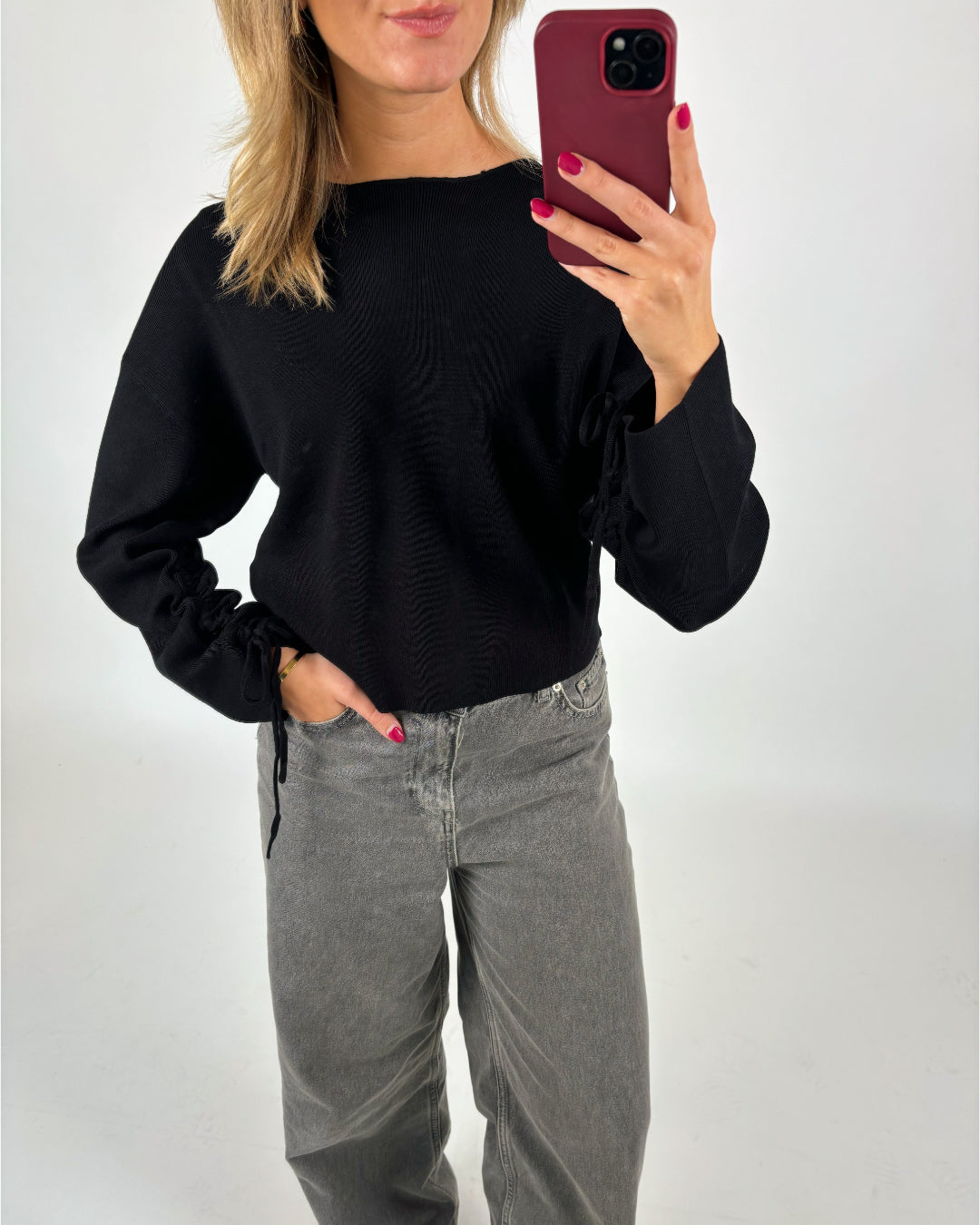 En person i sort trøje og grå jeans tager et spejlbillede med en rød telefon. Personen har lyse, løsthængende hår og rød neglelak. Baggrunden er hvid.