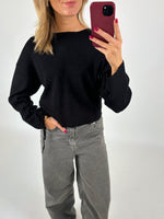 En person i sort trøje og grå jeans tager et spejlbillede med en rød telefon. Personen har lyse, løsthængende hår og rød neglelak. Baggrunden er hvid.