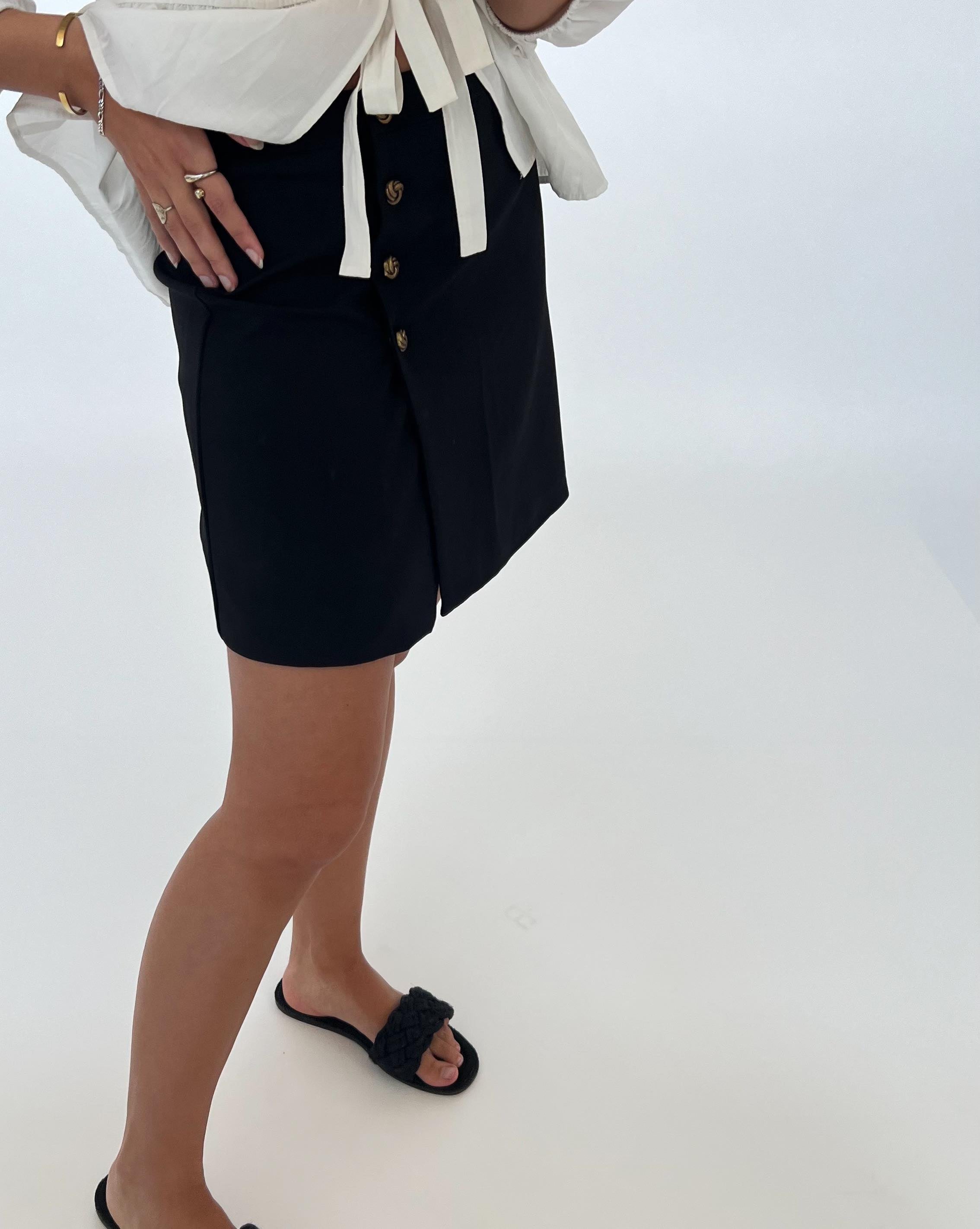 En person iført hvid skjorte og sort nederdel med knapper står i en lys, minimalistisk indendørs opsætning. Personen har sorte sandaler og smykker på.