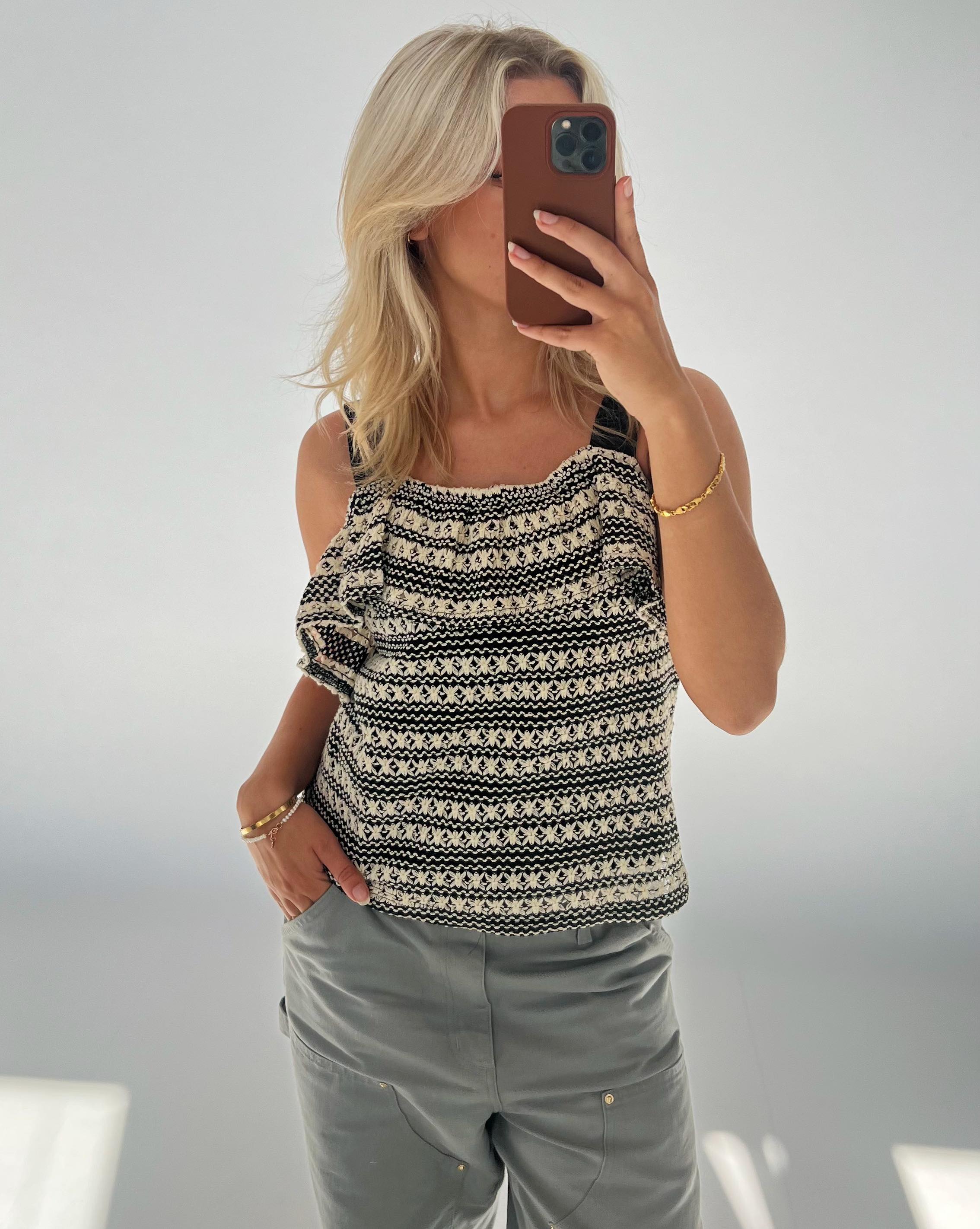 En person tager en selfie med en smartphone. Personen har en mønstret top på og grå bukser. Baggrunden er enkel og lys, hvilket fremhæver personen i fokus.