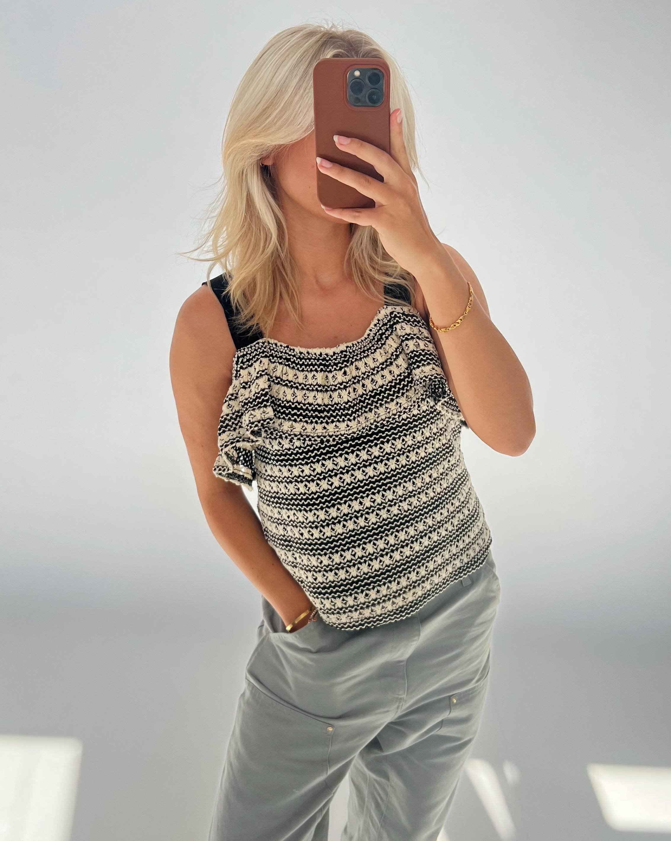En person tager en selfie med en smartphone, iført en sort-hvid mønstret top og grå bukser, i et lyst rum. Håret er lyst, og ansigtet er delvis skjult.