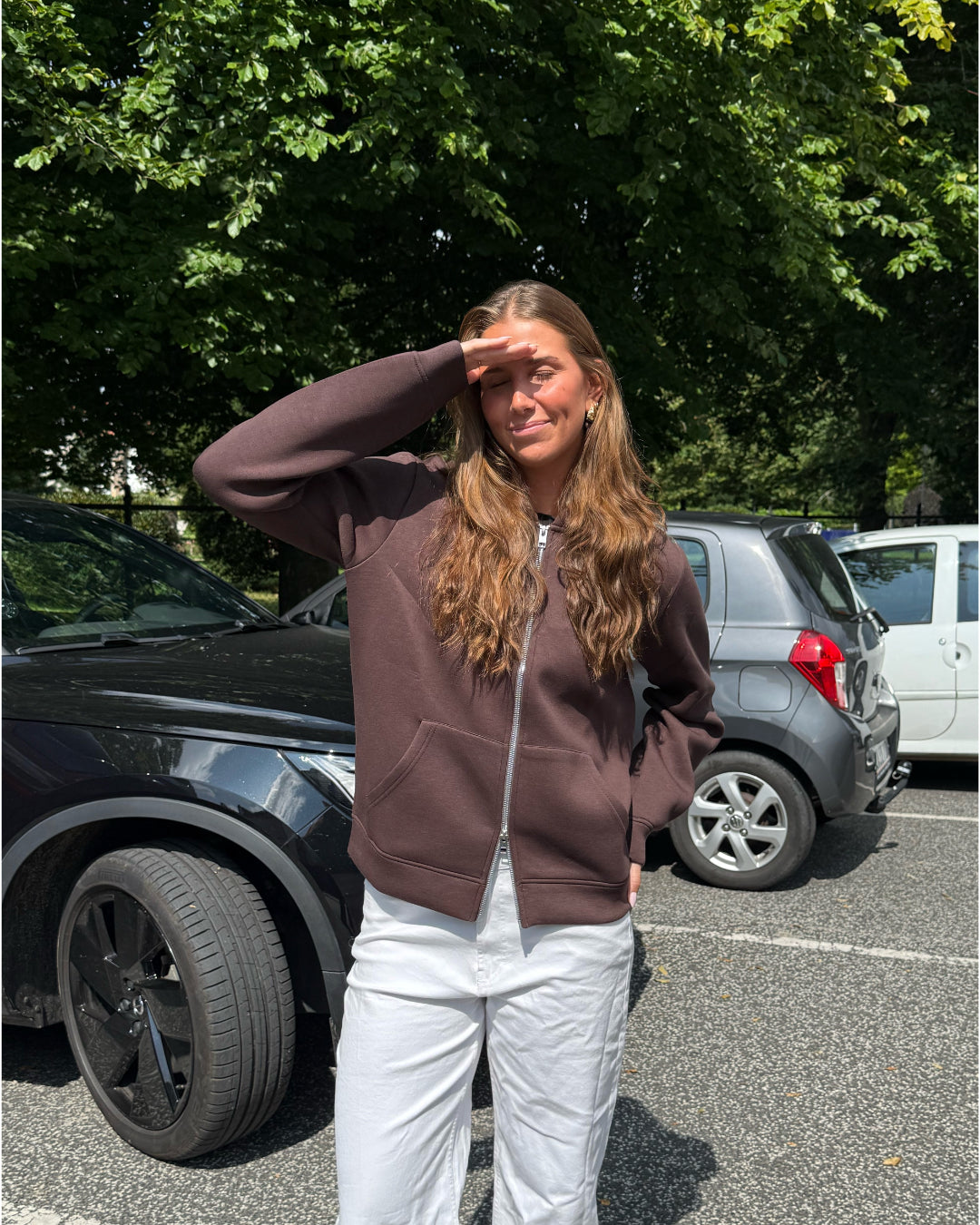Modelfoto af pige, som bærer EMMA hoodie, brun og NANNA jeans, hvid - alt fra BySofieSønderby. Baggrund er p-plads og træer