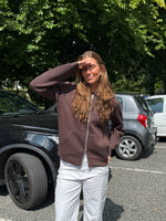 Modelfoto af pige, som bærer EMMA hoodie, brun og NANNA jeans, hvid - alt fra BySofieSønderby. Baggrund er p-plads og træer