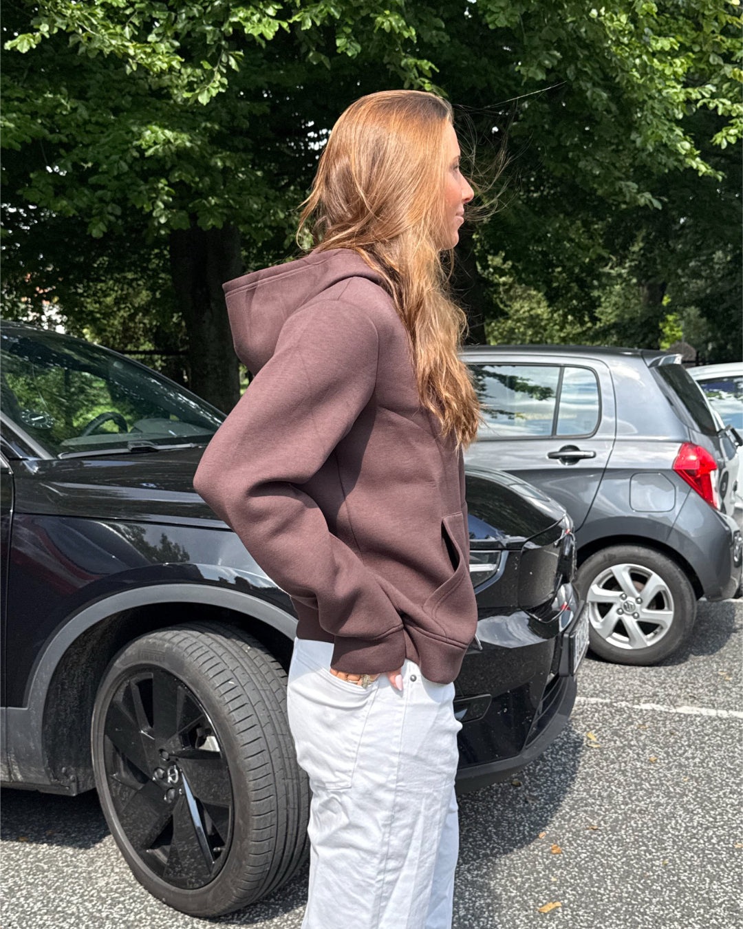 Modelfoto af pige, som bærer EMMA hoodie, brun og NANNA jeans, hvid - alt fra BySofieSønderby. Baggrund er p-plads og træer