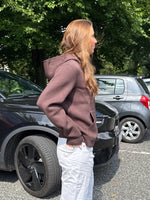 Modelfoto af pige, som bærer EMMA hoodie, brun og NANNA jeans, hvid - alt fra BySofieSønderby. Baggrund er p-plads og træer