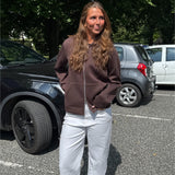 Modelfoto af pige, som bærer EMMA hoodie, brun og NANNA jeans, hvid - alt fra BySofieSønderby. Baggrund er p-plads og træer