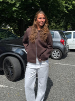 Modelfoto af pige, som bærer EMMA hoodie, brun og NANNA jeans, hvid - alt fra BySofieSønderby. Baggrund er p-plads og træer