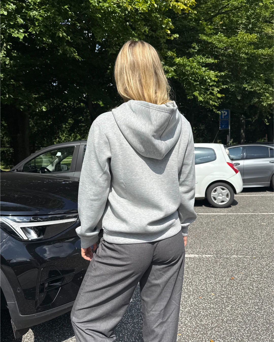 Modelfoto af pige, som bærer EMMA hoodie, lysegrå og MILAN bukser, grå - alt fra BySofieSønderby. Baggrund er p-plads og træer