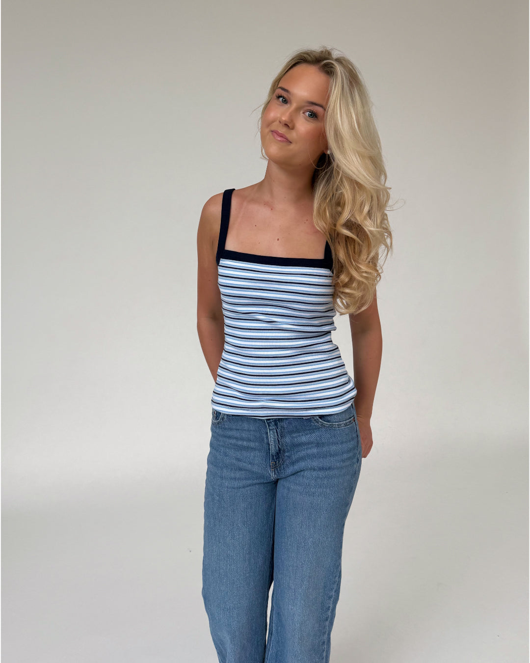 Kvinde står afslappet i afslappet tøj, iført en blå- og hvidstribet top og jeans, med hænderne bag ryggen i et neutralt rum.
